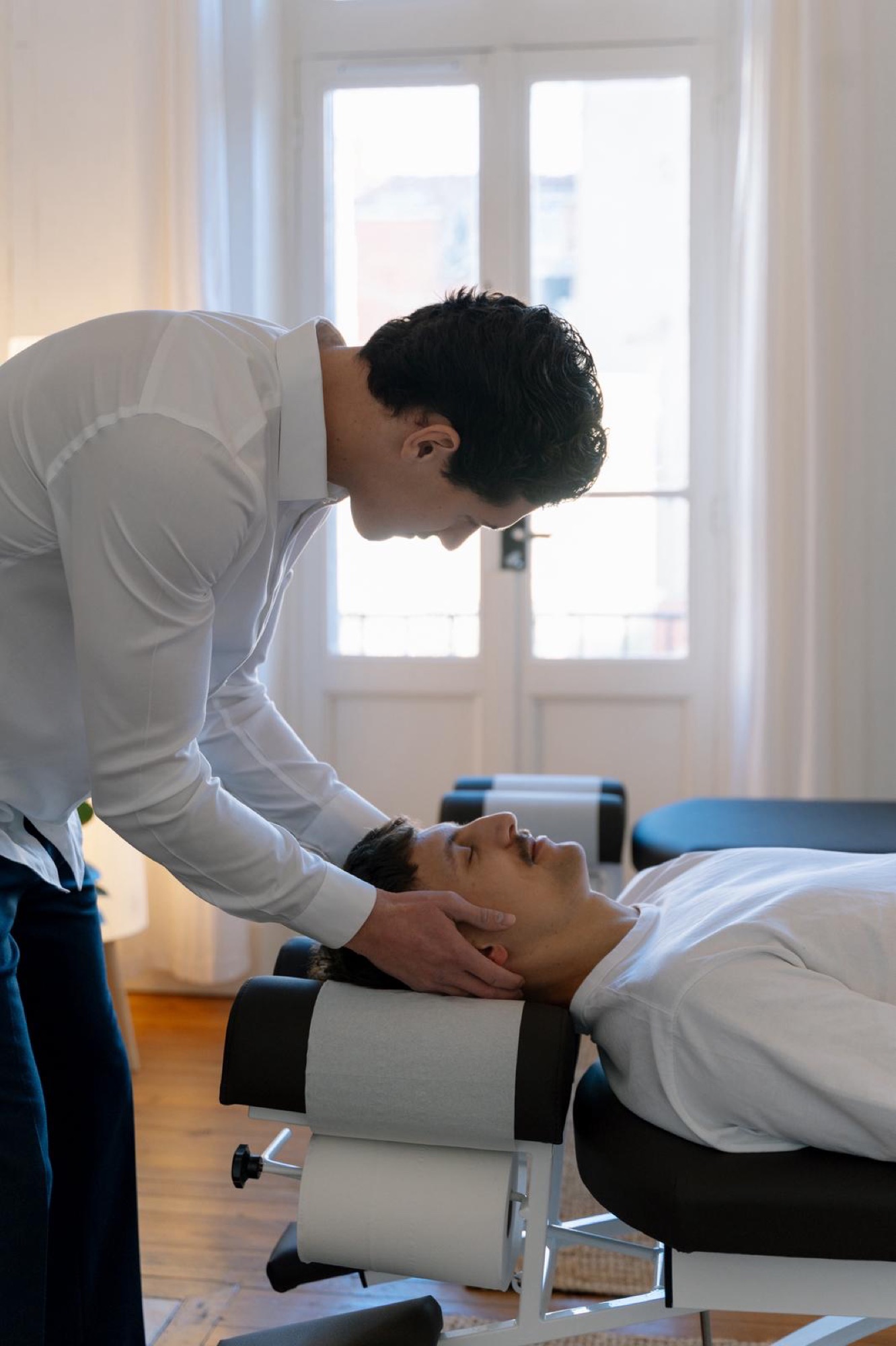 Thomas Nunes réalise un ajustement cervical à un patient allongé, devant la fenêtre du cabinet